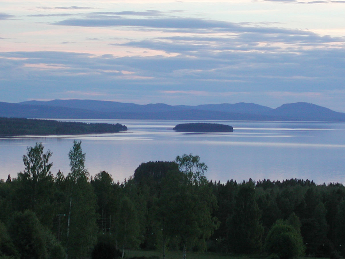View of a calm lake with a small wooded island, surrounded by forested shoreline and distant hills under a cloudy sky at dusk.