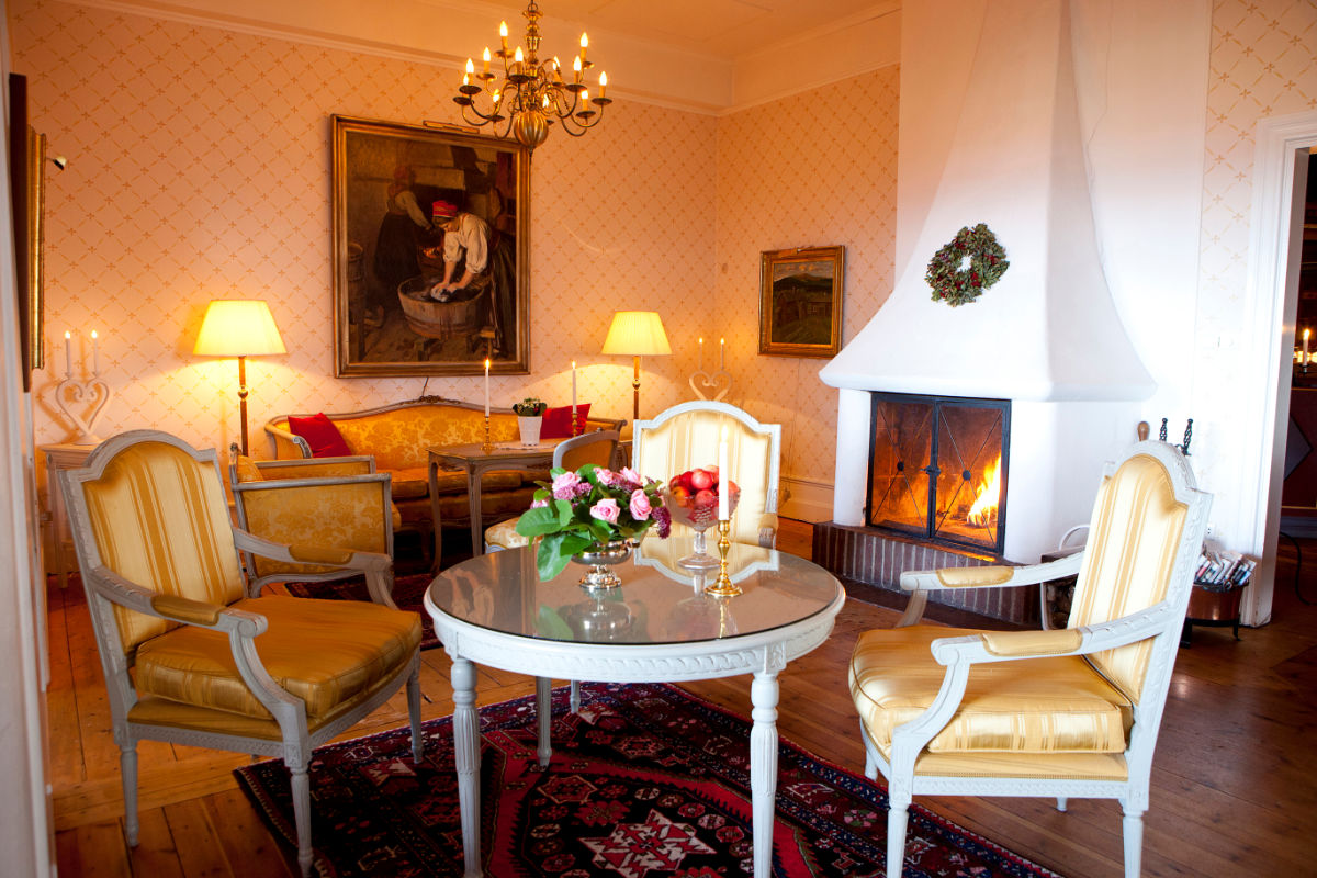 A cozy living room with yellow upholstered chairs, a round table with flowers, a lit fireplace, a chandelier, and paintings on patterned wallpapered walls.