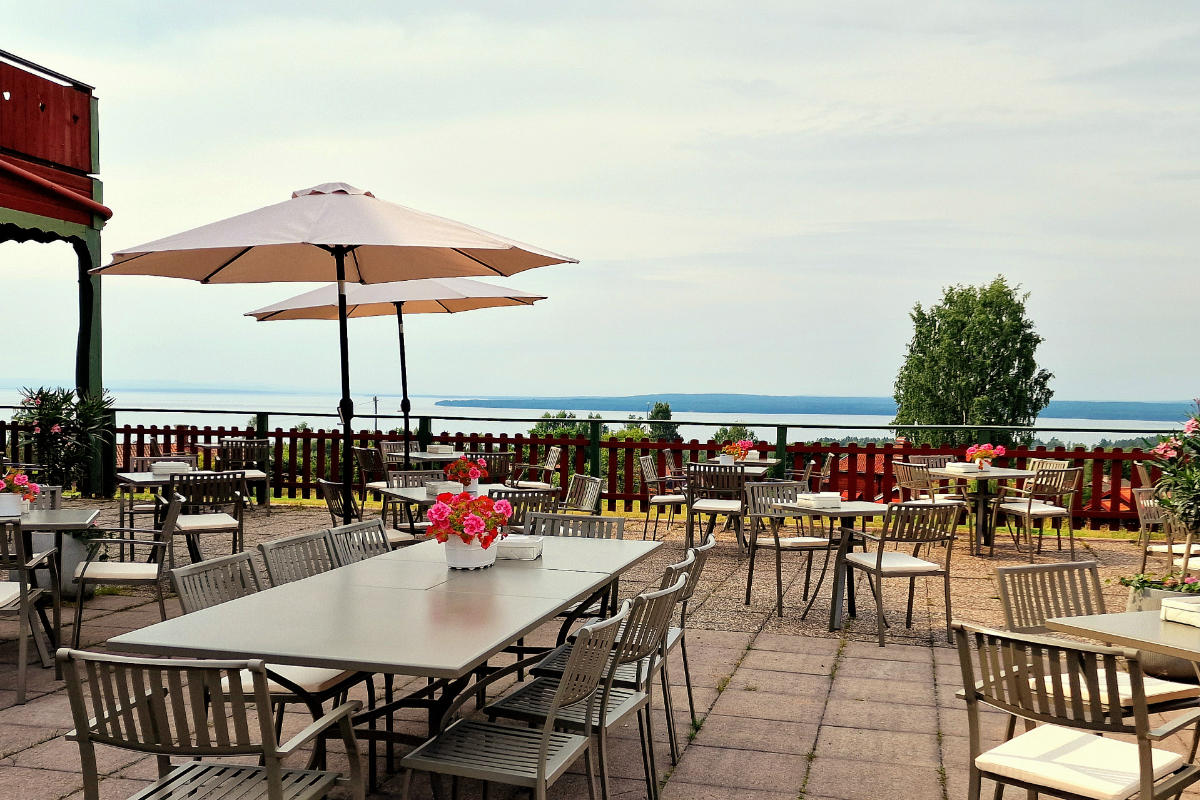 Outdoor patio with tables and chairs, some with flower centerpieces, overlooking a lake and distant shoreline under a partly cloudy sky.