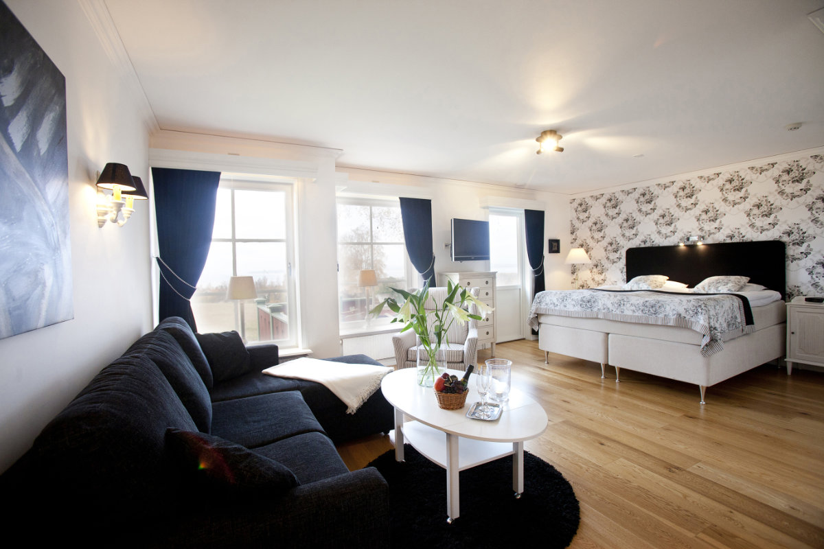 A bright, modern hotel room features a king-sized bed, a black sofa, a white coffee table with plants, large windows, and floral wallpaper accent.