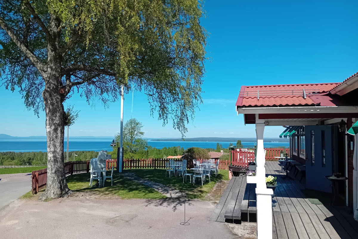 Outdoor seating area by a tree with white tables and chairs, overlooking a blue lake under a clear sky, next to a red-roofed building.