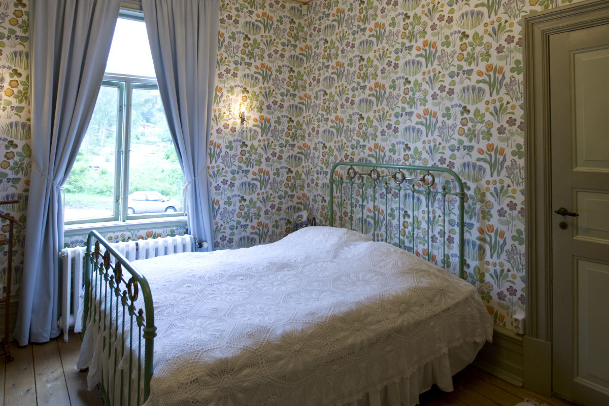 A small bedroom at Villa Sjötorp with floral wallpaper, a metal bed frame, white bedding, light blue curtains, a window, and a closed door.