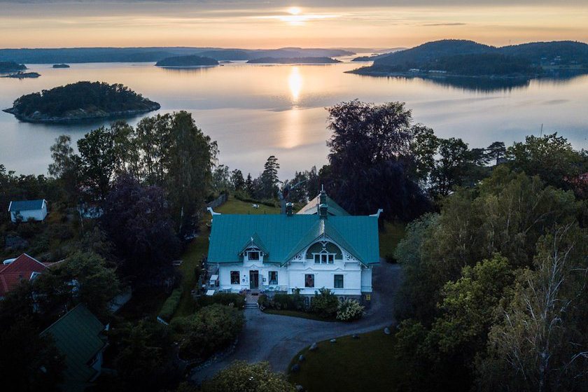 A large white house, Villa Sjötorp, with a green roof stands by a calm lake at sunset, with small forested islands and distant hills visible across the water.