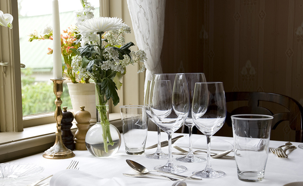 A set dining table at Villa Sjötorp features wine glasses, water glasses, cutlery, candlesticks, and a vase of flowers by a window with light curtains.