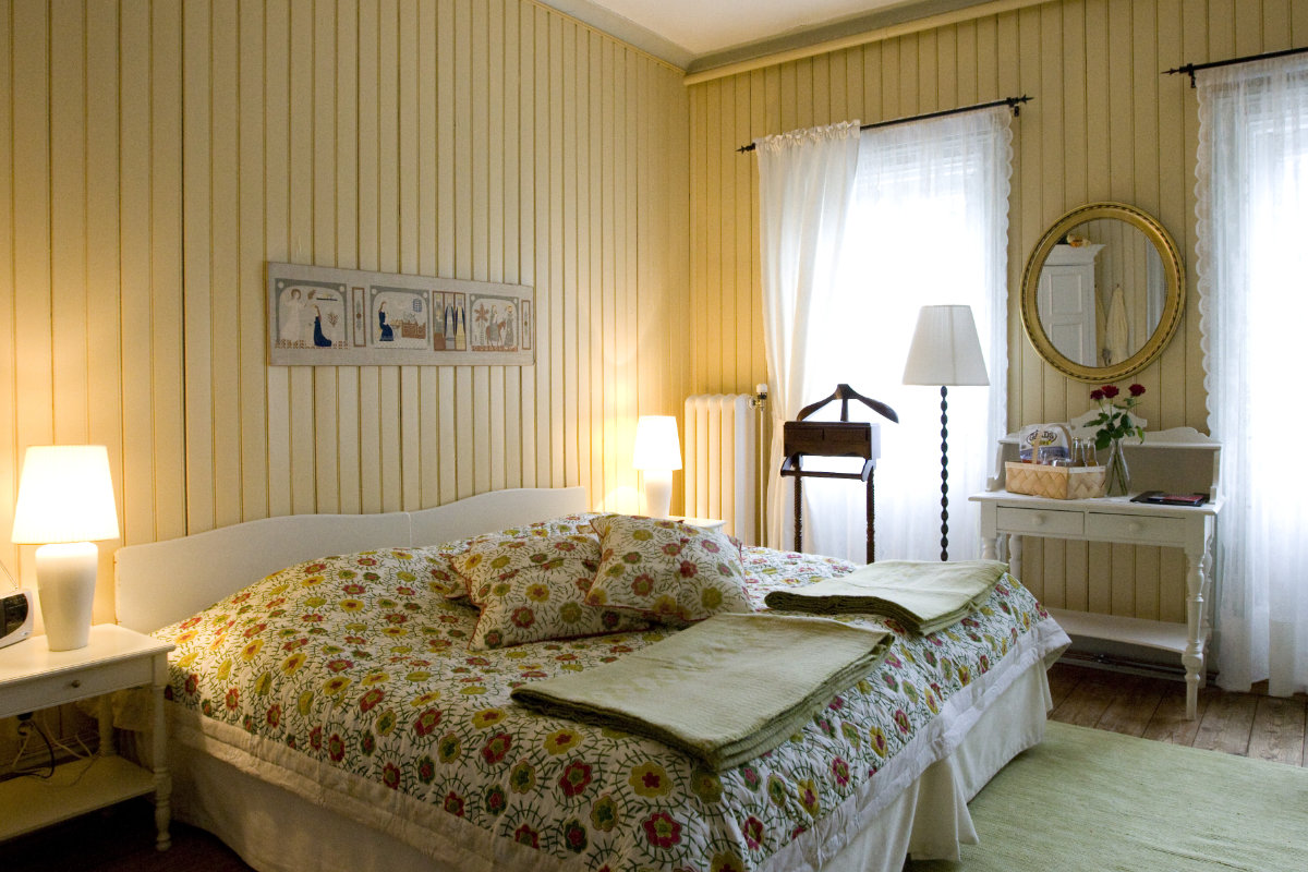 A cozy bedroom at Villa Sjötorp with a double bed, floral bedding, two bedside tables with lamps, a mirror above a desk, and two curtained windows letting in natural light.
