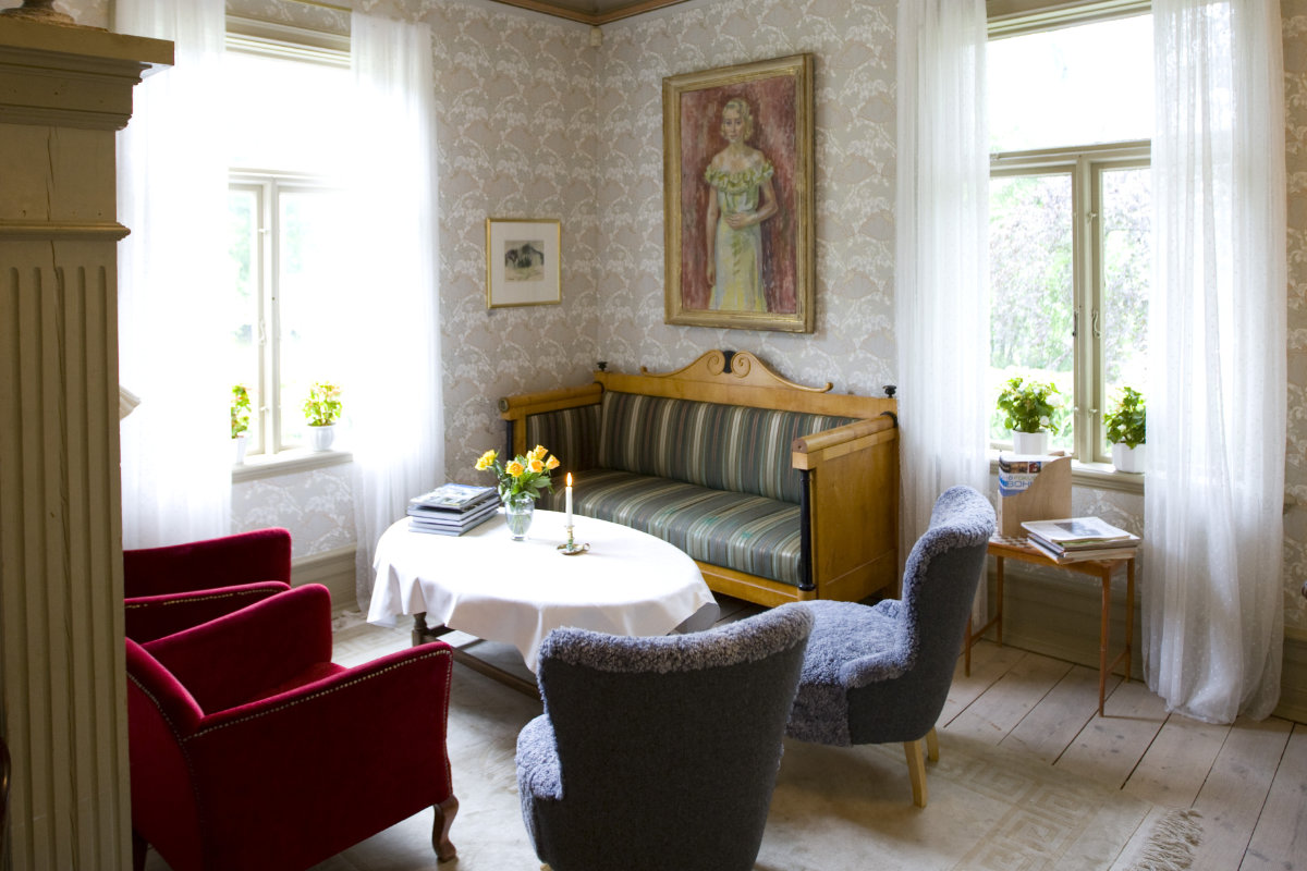 A bright living room at Villa Sjötorp with two gray armchairs, a red armchair, a striped wooden sofa, a round table with flowers, and two large windows with sheer curtains.