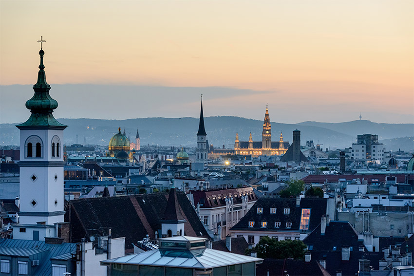 7 vacaciones en Europa que podrían cambiar su vida 2 Vista del paisaje urbano de Viena al atardecer, con campanarios de iglesias, tejados históricos y edificios iluminados con colinas al fondo.