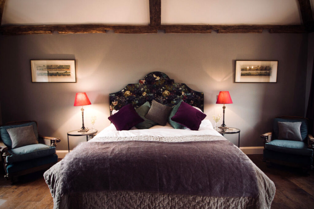 Escapadas otoñales: Explore los parques nacionales europeos a todo color 12 Una cama pulcramente hecha con un cabecero de flores, una manta púrpura y almohadas variadas, flanqueada por dos sillones y lámparas rojas, en un dormitorio acogedor y suavemente iluminado.