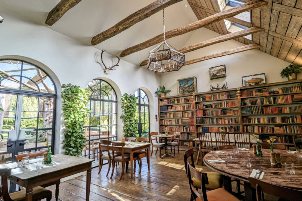 De nouvelles façons de voir le monde en 2026 11 Une salle à manger ensoleillée avec des poutres en bois, des fenêtres cintrées, des plantes en pot, un mur tapissé de livres et plusieurs tables en bois dressées pour un repas.