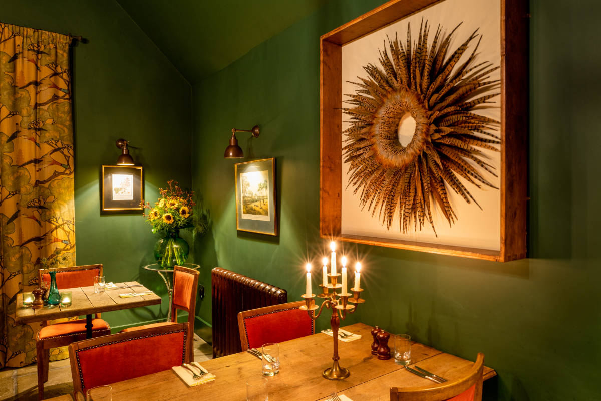 Une salle de restaurant chaleureusement éclairée avec des murs verts, des chaises rouges, des tables en bois et une grande œuvre d'art décorative en forme de soleil suspendue au-dessus d'un candélabre.