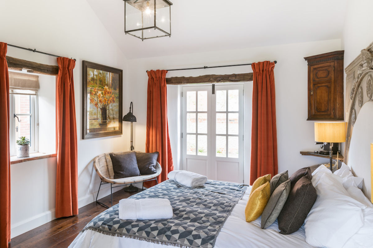 Chambre à coucher lumineuse avec un lit double, un édredon à motifs, des coussins assortis, des rideaux orange, un plancher en bois et une chaise près d'une fenêtre. Des portes-fenêtres et des œuvres d'art encadrées décorent la pièce.