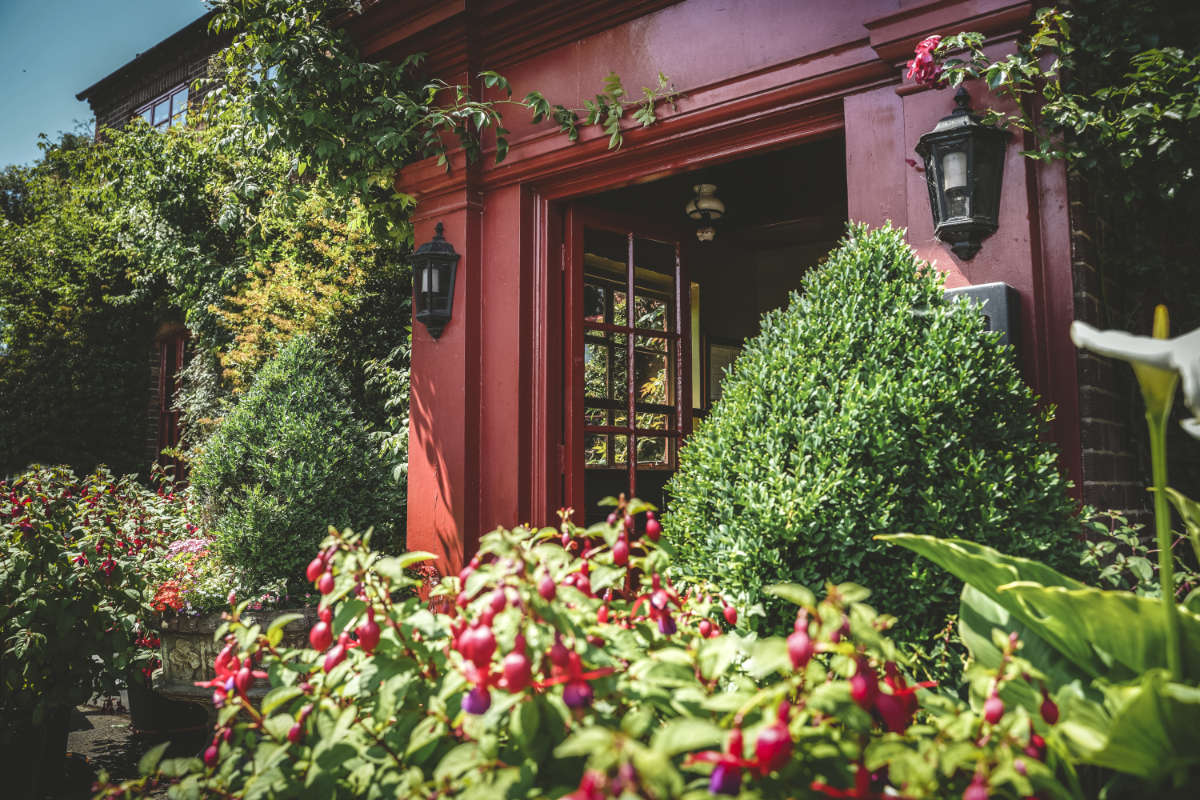 Una puerta roja con farolillos negros está rodeada de arbustos verdes y plantas en flor delante de un edificio de ladrillo.
