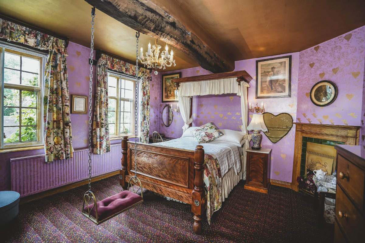 Un dormitorio con paredes lilas, cortinas de flores, una cama de madera con dosel, lámpara de araña, cuadros de arte y un columpio acolchado colgado del techo cerca de las ventanas.