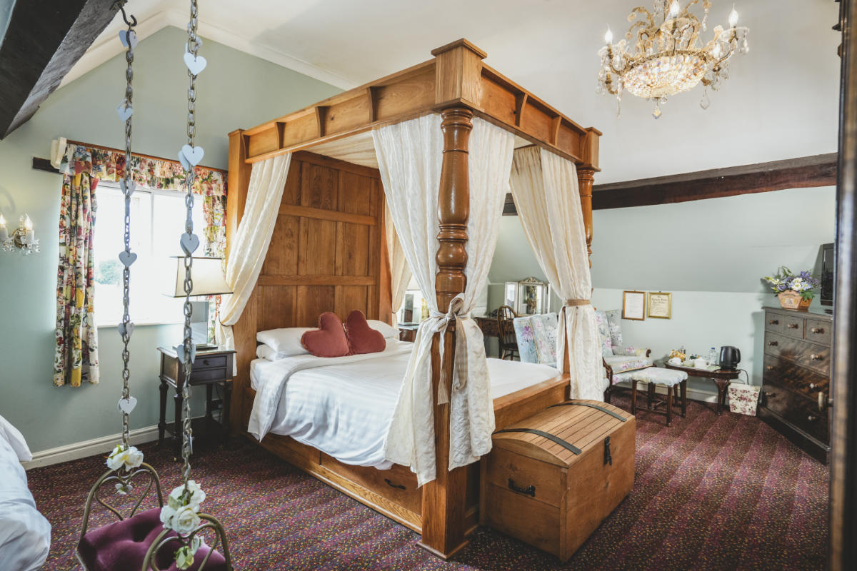 Un acogedor dormitorio con una cama de madera con dosel, cortinas blancas, visillos florales, lámpara de araña y muebles clásicos sobre una alfombra estampada.