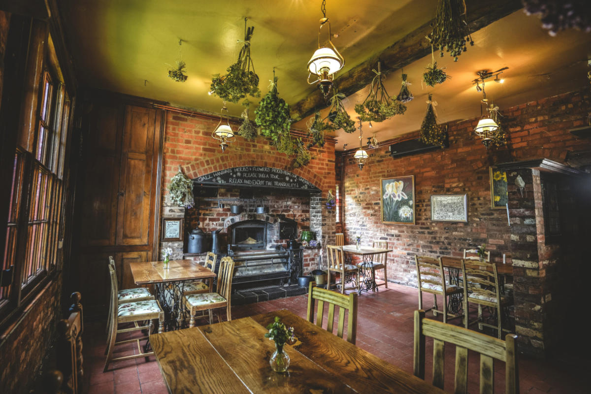 Acogedor comedor rústico con mesas y sillas de madera, paredes de ladrillo visto, hierbas secas colgantes y una gran chimenea horno. La cálida iluminación crea un ambiente acogedor.