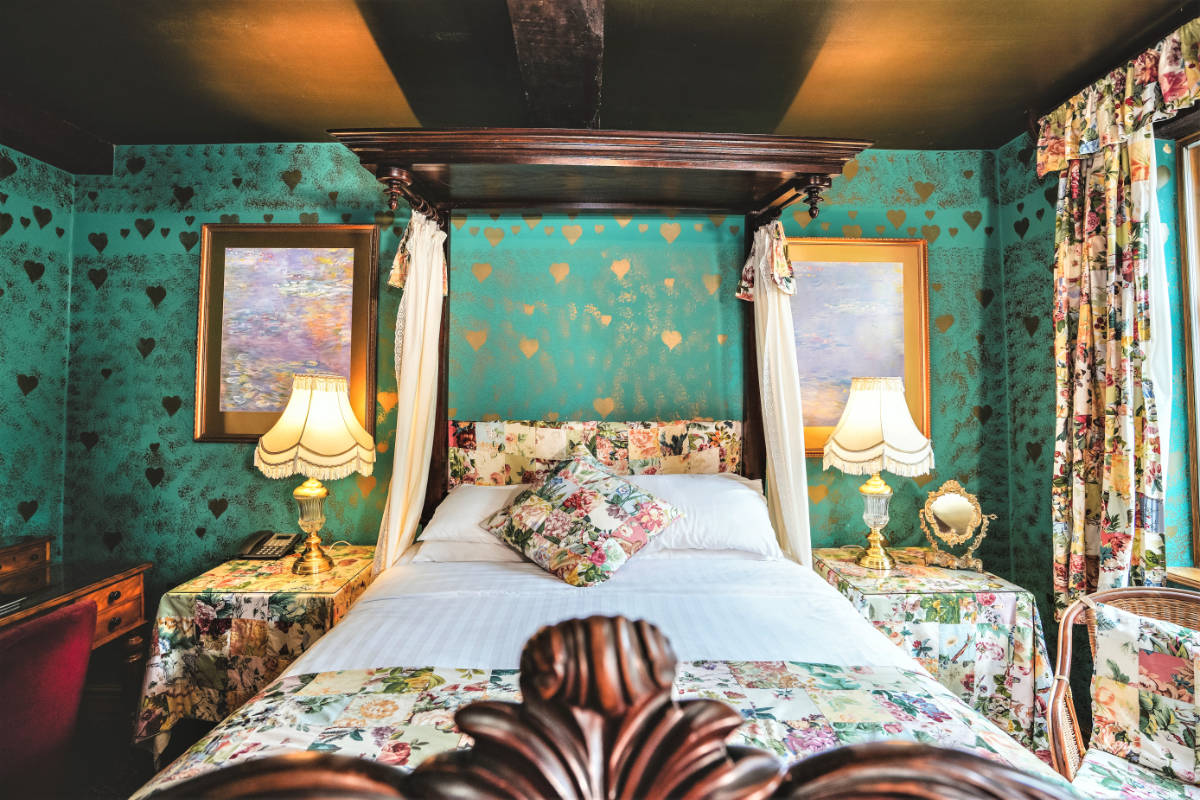 Un acogedor dormitorio con cama con dosel, ropa de cama de flores, papel pintado estampado con corazones dorados, dos mesillas de noche con lámparas y cuadros en las paredes.