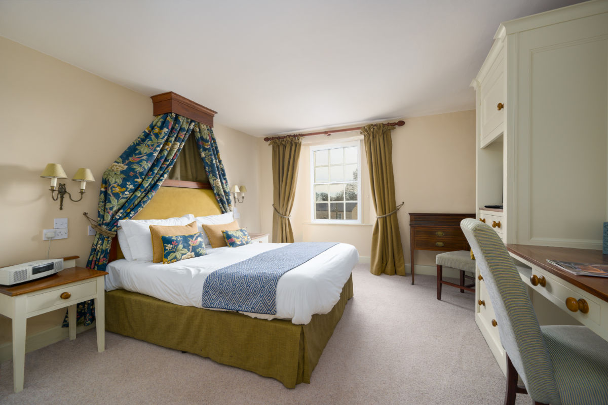 En una luminosa habitación de hotel con paredes beige, un escritorio, una silla y una ventana con cortinas verdes, hay una cama bien hecha con un dosel azul y amarillo.