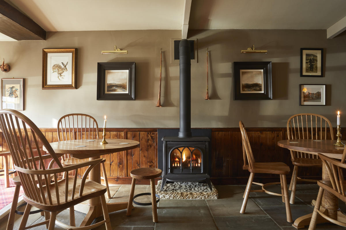 Una acogedora sala con sillas de madera y mesas redondas, una estufa de leña encendida en el centro y obras de arte enmarcadas en las paredes. En las mesas hay velas.