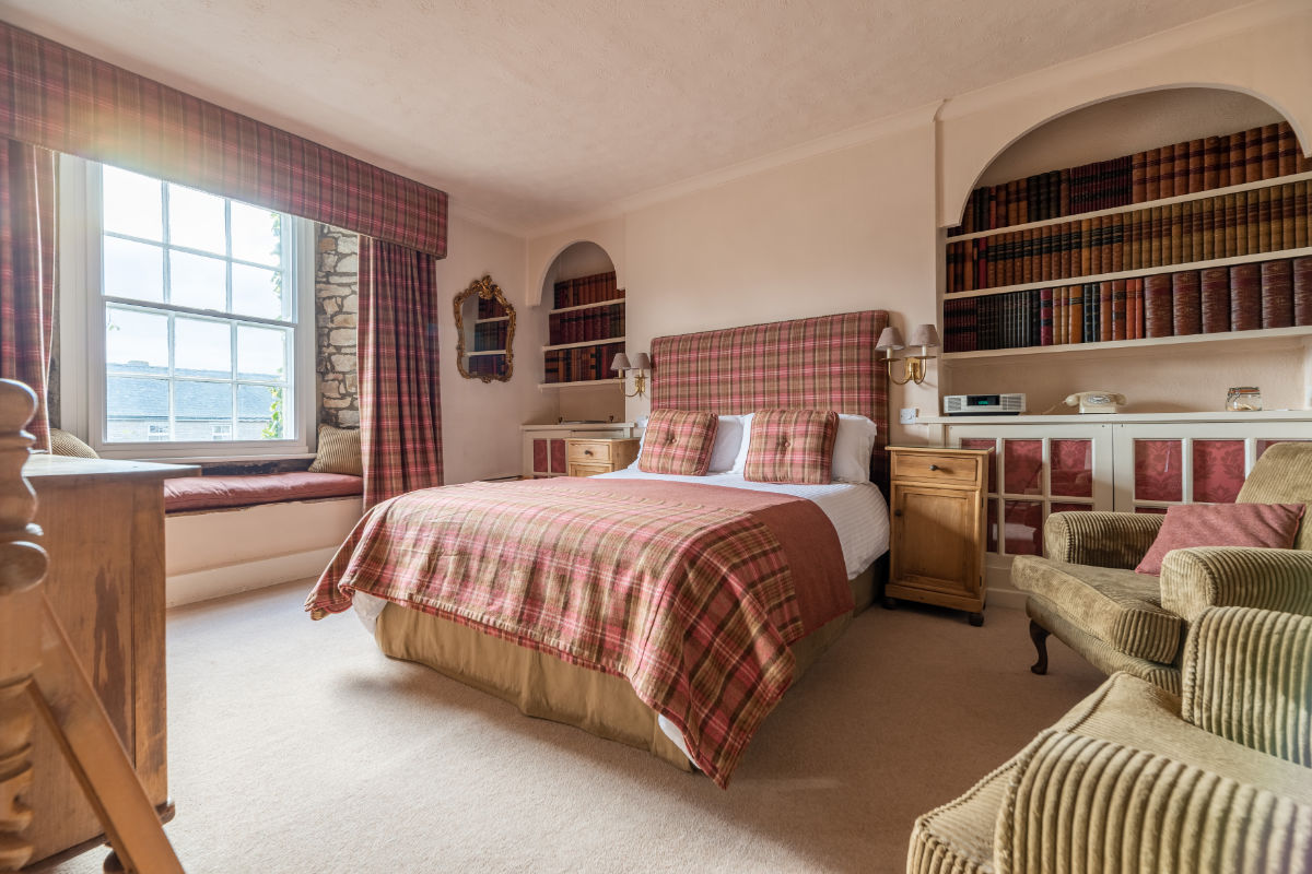 Un acogedor dormitorio con cama de matrimonio, ropa de cama a cuadros, sillón, estanterías empotradas llenas de libros, un escritorio de madera y un asiento en la ventana con vistas al exterior.