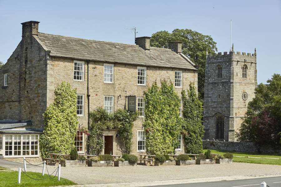 Un bâtiment historique en pierre couvert de lierre se trouve à côté d'une église en pierre avec une tour et une horloge, le tout par une journée ensoleillée avec un ciel clair.