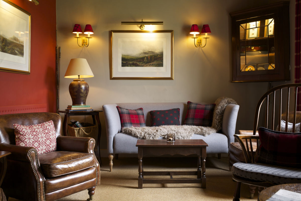 Un acogedor salón con un sofá gris, cojines de cuadros escoceses, sillón de cuero, mesa de madera, obras de arte en la pared y una cálida iluminación de apliques y una lámpara de sobremesa.
