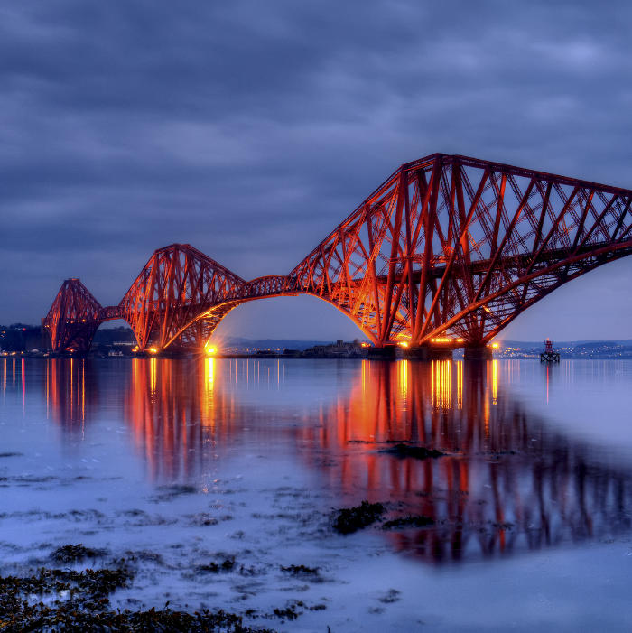 Il Forth Bridge, un ponte ferroviario rosso a sbalzo, è illuminato al crepuscolo e si riflette nell'acqua calma sotto un cielo nuvoloso.