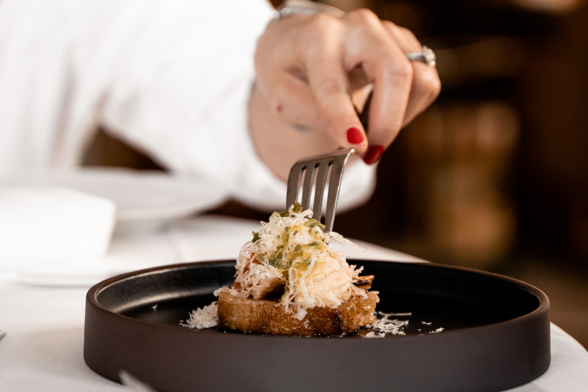 Une personne saisit à l'aide d'une fourchette un morceau de pain grillé garni de fromage râpé et d'herbes, servi dans une assiette noire.