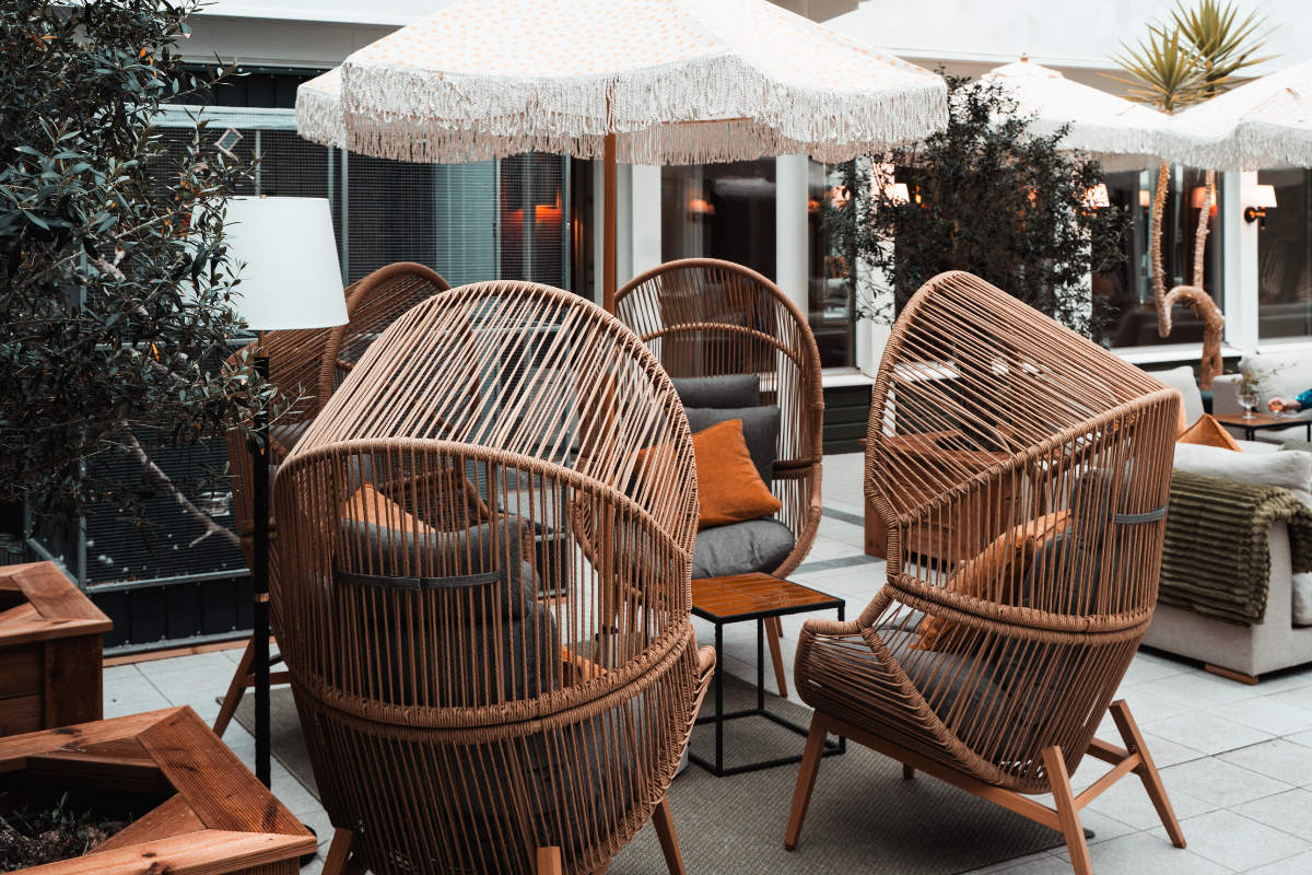 Quatre chaises longues en rotin avec coussins entourent une petite table sur un patio, sous de grands parasols blancs à franges, avec des plantes et des décorations d'extérieur en arrière-plan.