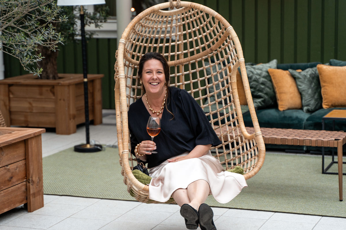 Une femme est assise dans un fauteuil suspendu en osier, tenant un verre de vin rosé et souriant. Des coussins verts et des meubles en bois se trouvent à l'arrière-plan.