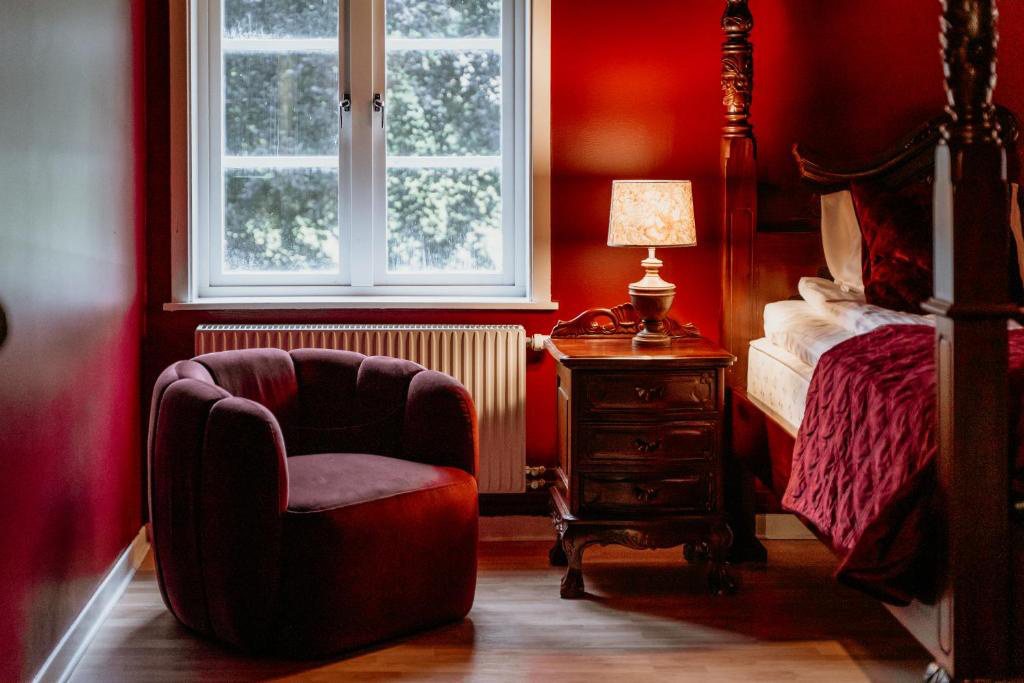 Une chambre aux murs rouge foncé, un fauteuil rouge en peluche, un lit en bois orné, une table de nuit avec une lampe et une fenêtre laissant entrer la lumière du jour.