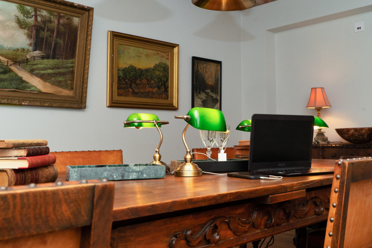 Ein Holzschreibtisch mit einem Laptop, grün getönte Banker-Lampen, gestapelte Bücher und gerahmte Bilder an den Wänden in einem Büro im traditionellen Stil.