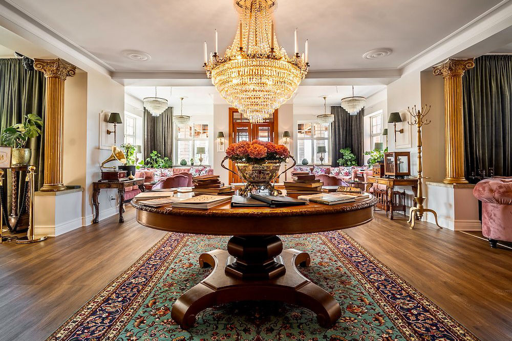 Soggiorno decorato con un grande lampadario, tavolo rotondo in legno con libri e fiori, tappeto a motivi, colonne dorate e sedute in tessuto sullo sfondo.