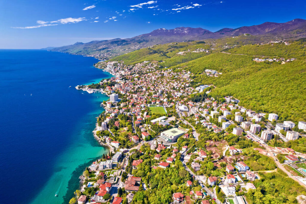 Aerial view of one of the best coastal towns for late summer, featuring numerous buildings, green hills, and a clear blue sea under a bright, sunny sky.