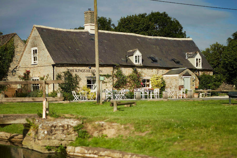 Stone building labeled "King's Head Inn" with outdoor seating, surrounded by grass and trees beneath a clear sky.