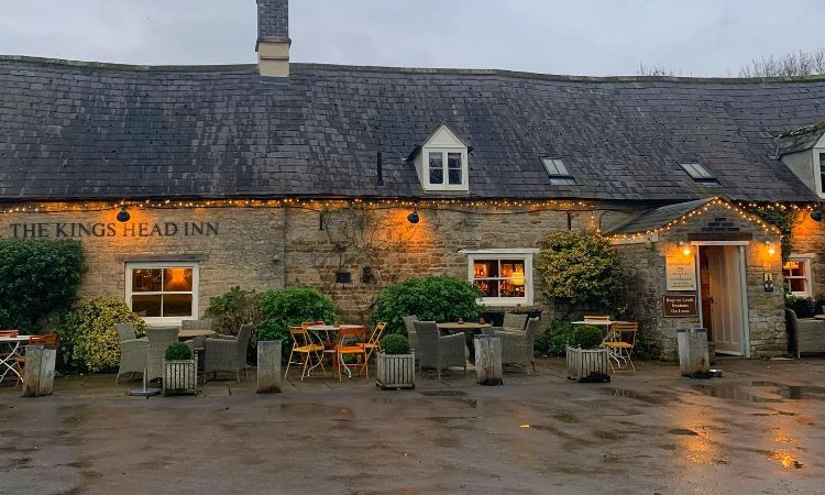 Stone inn with string lights, outdoor tables and chairs, and a sign reading "King's Head Inn" welcomes guests on a wet, overcast day.