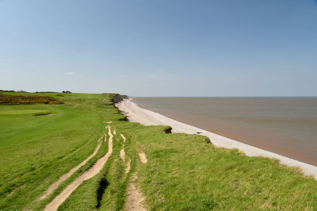 The Norfolk Coast path runs along a sloped cliff beside a narrow pebble beach and calm sea under a clear blue sky.