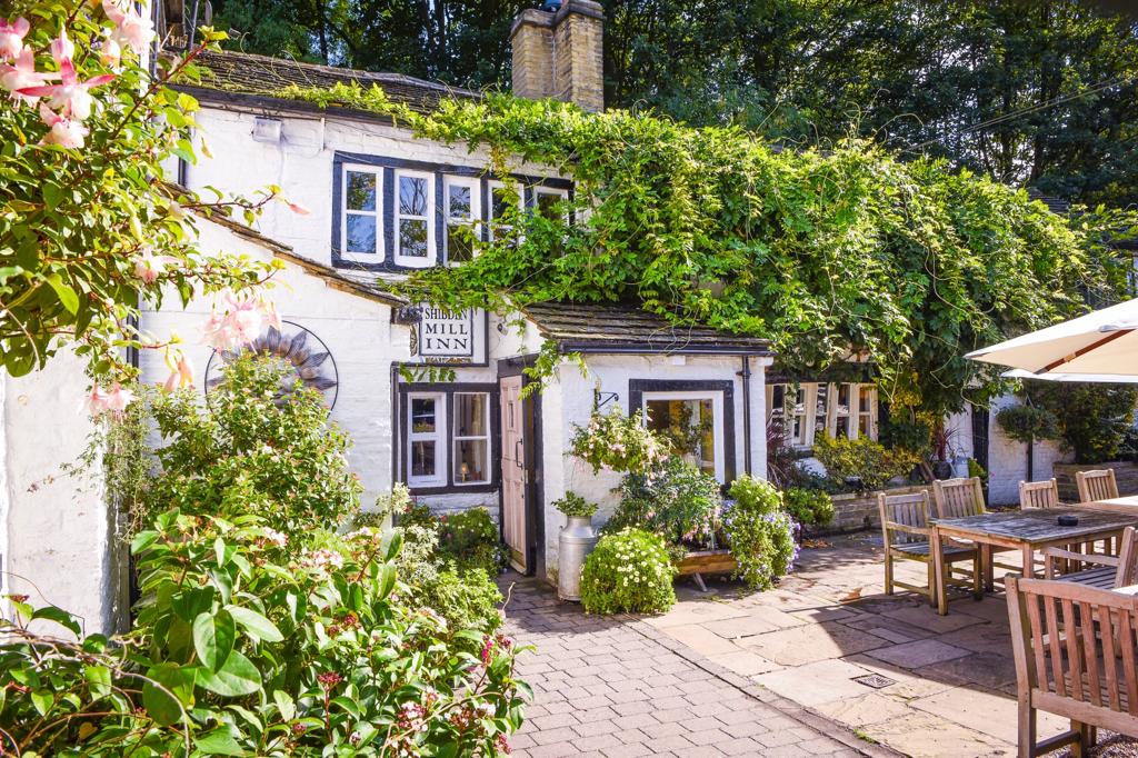 A white cottage with a sign reading "Mill Inn," reminiscent of Shibden Mill Inn, is covered in green vines, surrounded by plants, with wooden tables and chairs on a sunny patio.