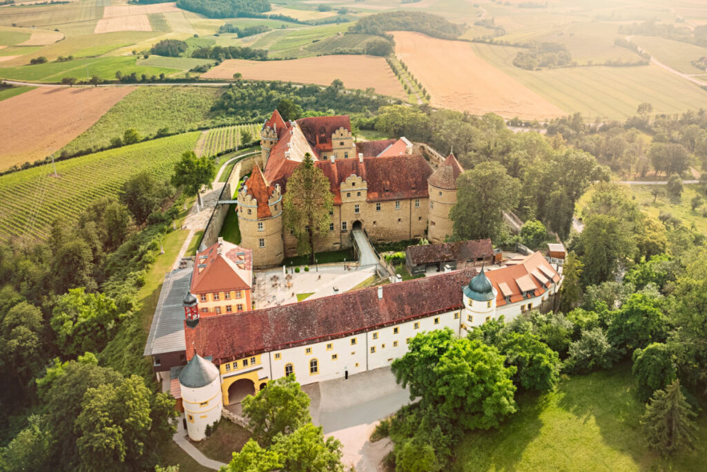Aerial view of a large historic castle complex with red-tiled roofs, surrounded by trees and fields in a rural landscape, showcasing the timeless beauty of castles in Europe.