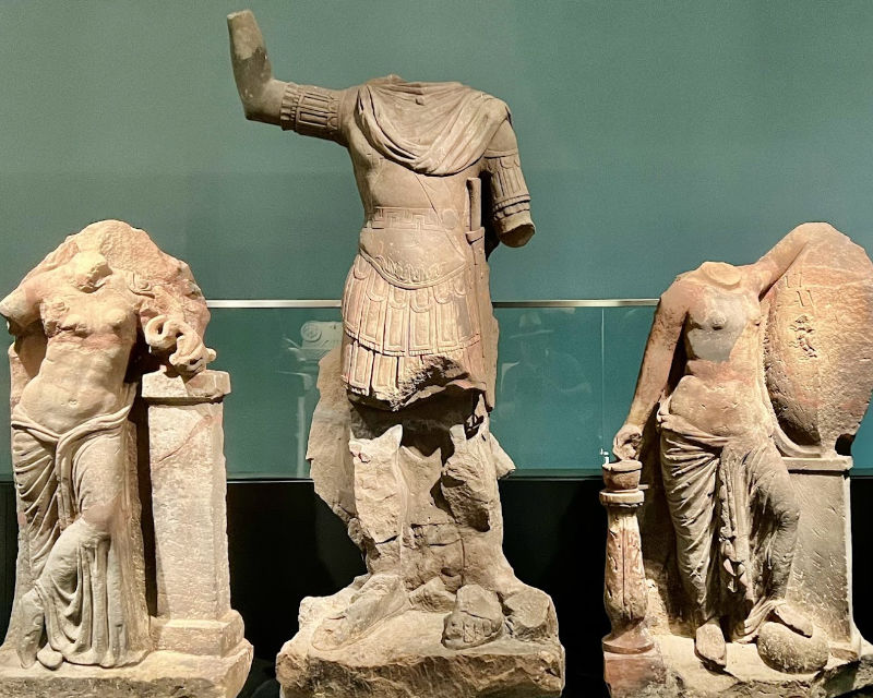 Drei kopflose antike Statuen - zwei drapierte weibliche Figuren, die eine gepanzerte männliche Figur mit einem erhobenen, gebrochenen Arm flankieren - sind im Römermuseum Weißenburg, Museum Bayern, vor einem schlichten Hintergrund ausgestellt.