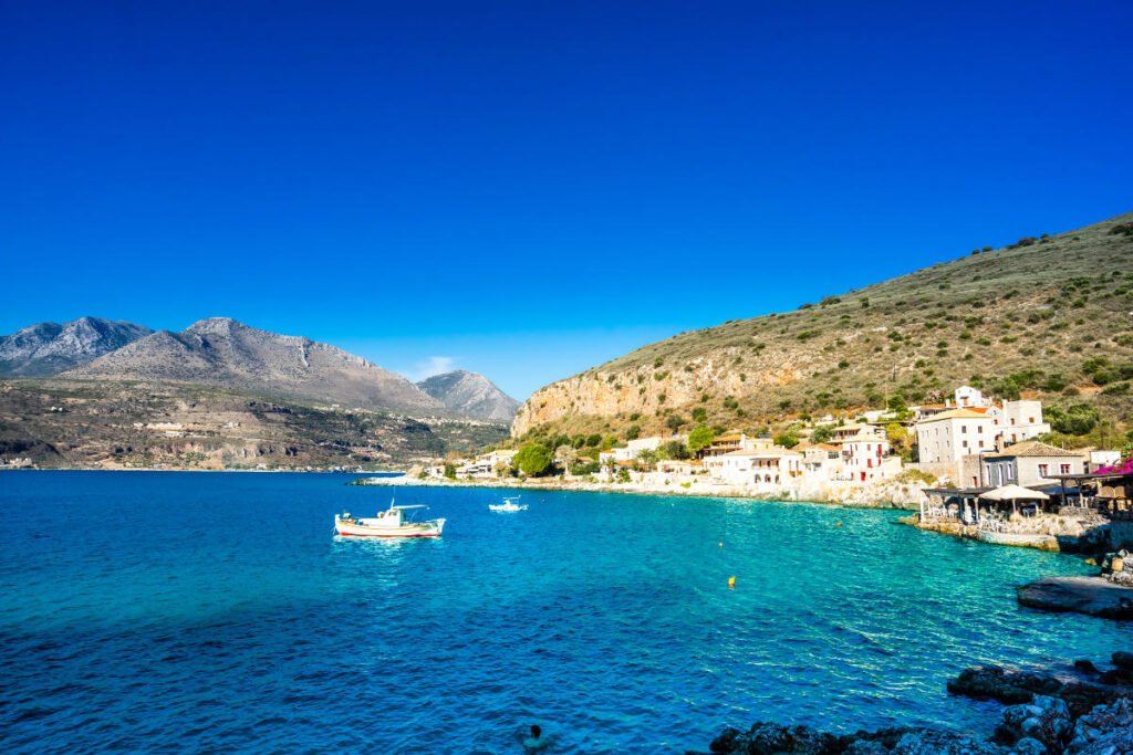 A coastal village with stone houses sits by a clear blue sea, perfect for those seeking coastal towns for late summer. Small boats float on the water, while green hills rise in the background under a bright blue sky.