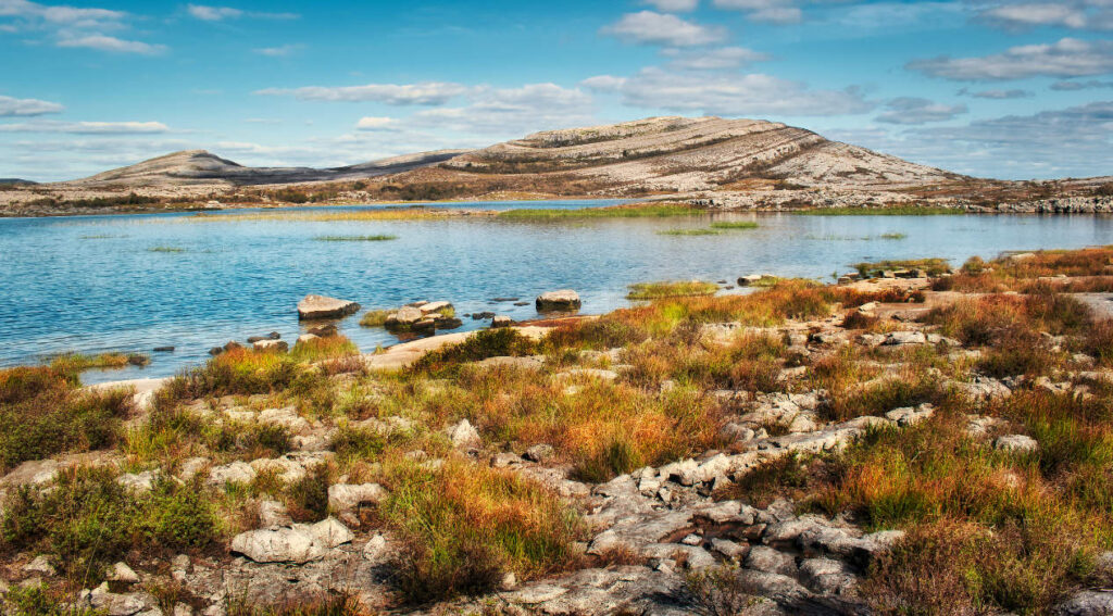 Rocky landscape with low vegetation in the foreground, a calm lake in the middle, and rounded hills under a partly cloudy sky in the background.