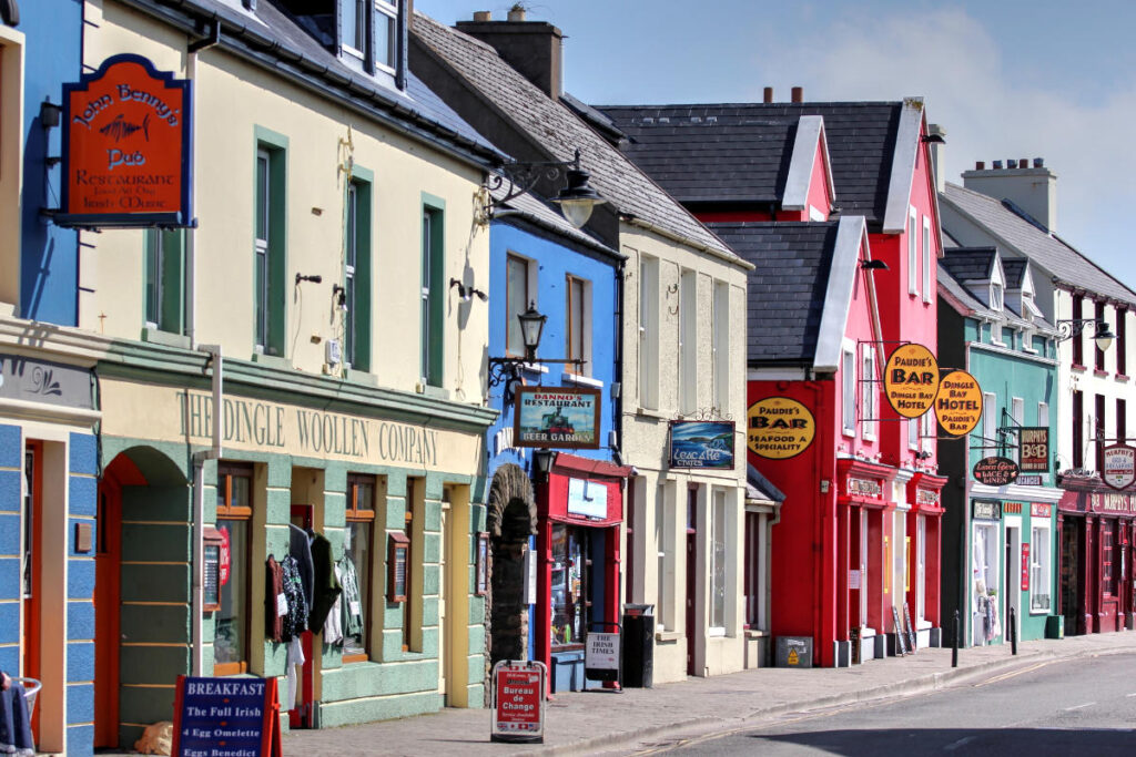 A row of colorful buildings with shops, pubs, and restaurants line a street in one of the charming coastal towns for late summer. Signs and hanging lights are visible on the inviting storefronts.