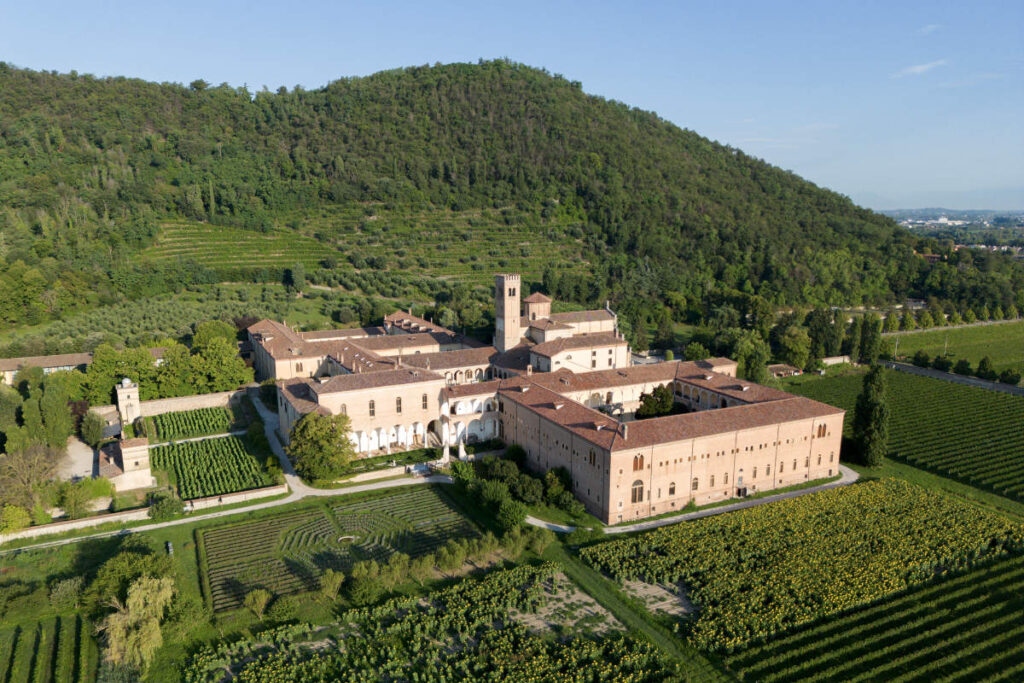 Aerial view of a large monastery complex with red-tiled roofs surrounded by vineyards and trees at the base of a green, forested hill.