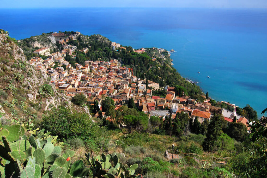 A coastal hillside town with clustered buildings and red rooftops overlooks the blue sea, surrounded by green vegetation and cacti—one of the perfect coastal towns for late summer escapes.
