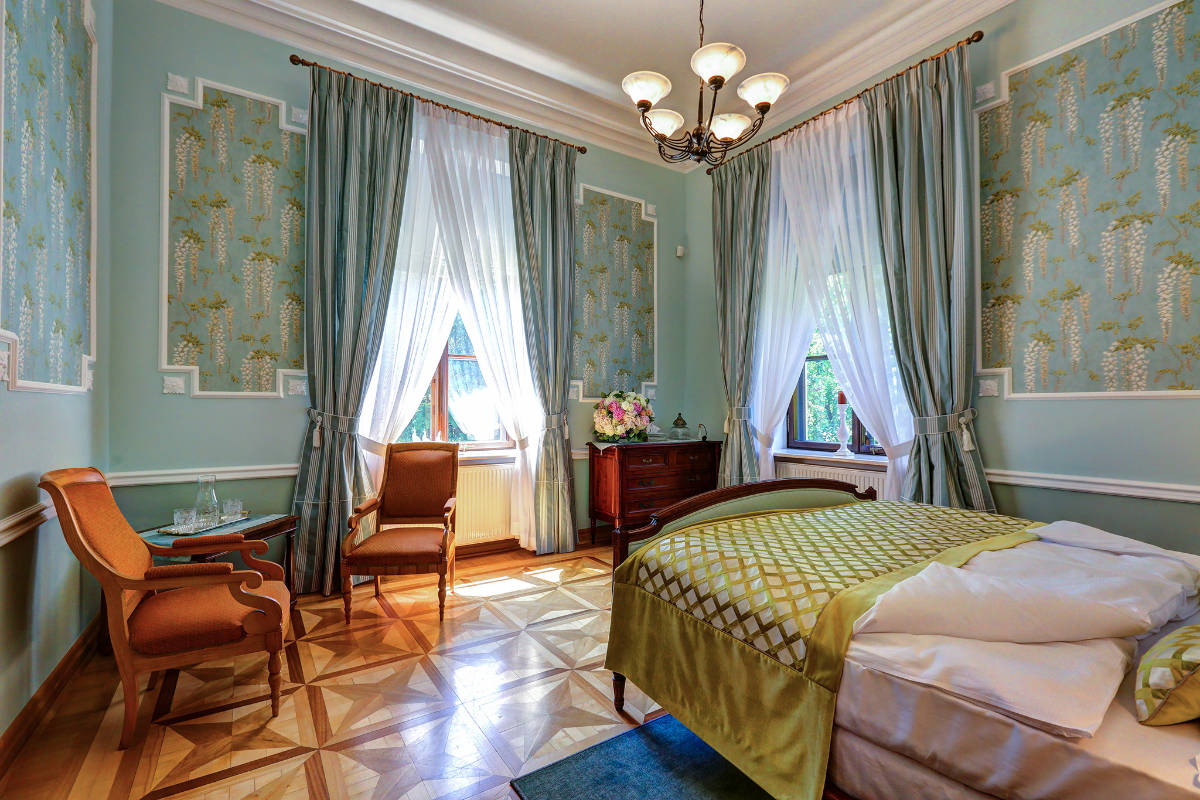 Elegant bedroom at Pałac Rozalin with patterned wallpaper, parquet flooring, a double bed, antique chairs, a wooden dresser, and large windows draped with sheer curtains.