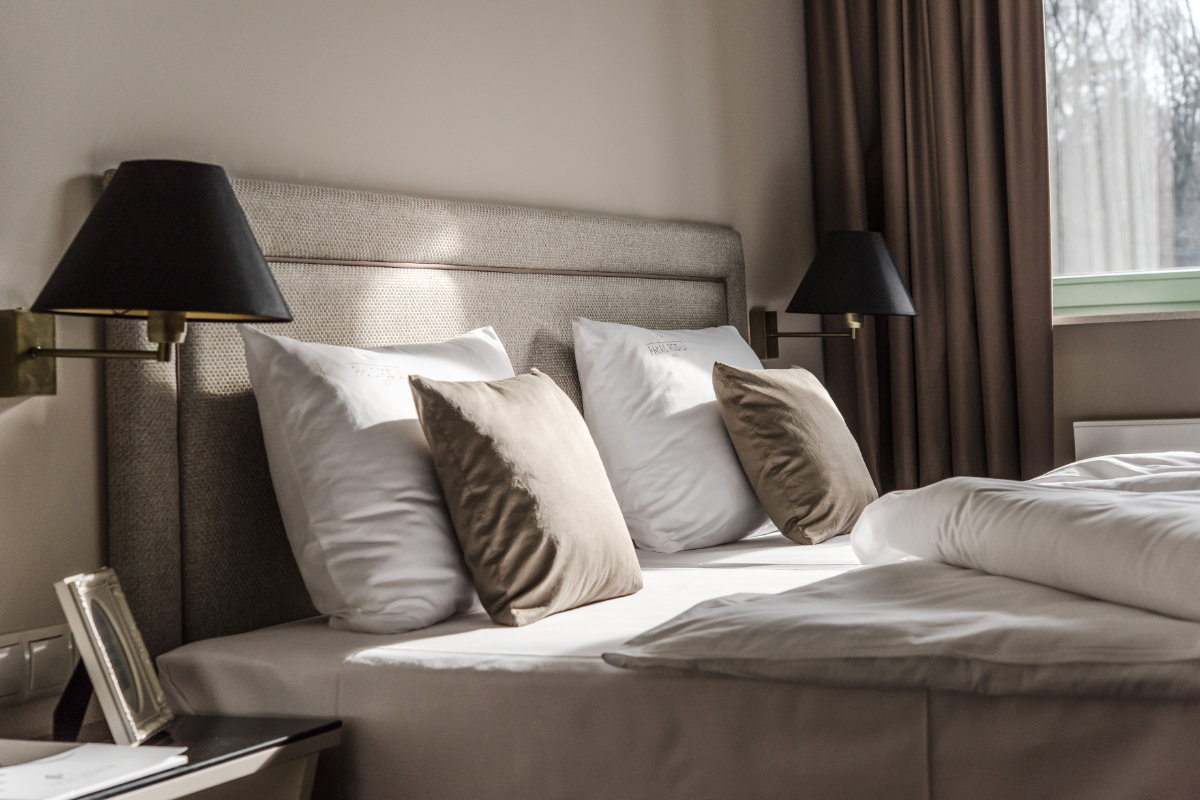 A neatly made hotel bed at Pałac Rozalin features white linens, four pillows, two brown cushions, two wall-mounted black lamps, and a window with closed brown curtains.