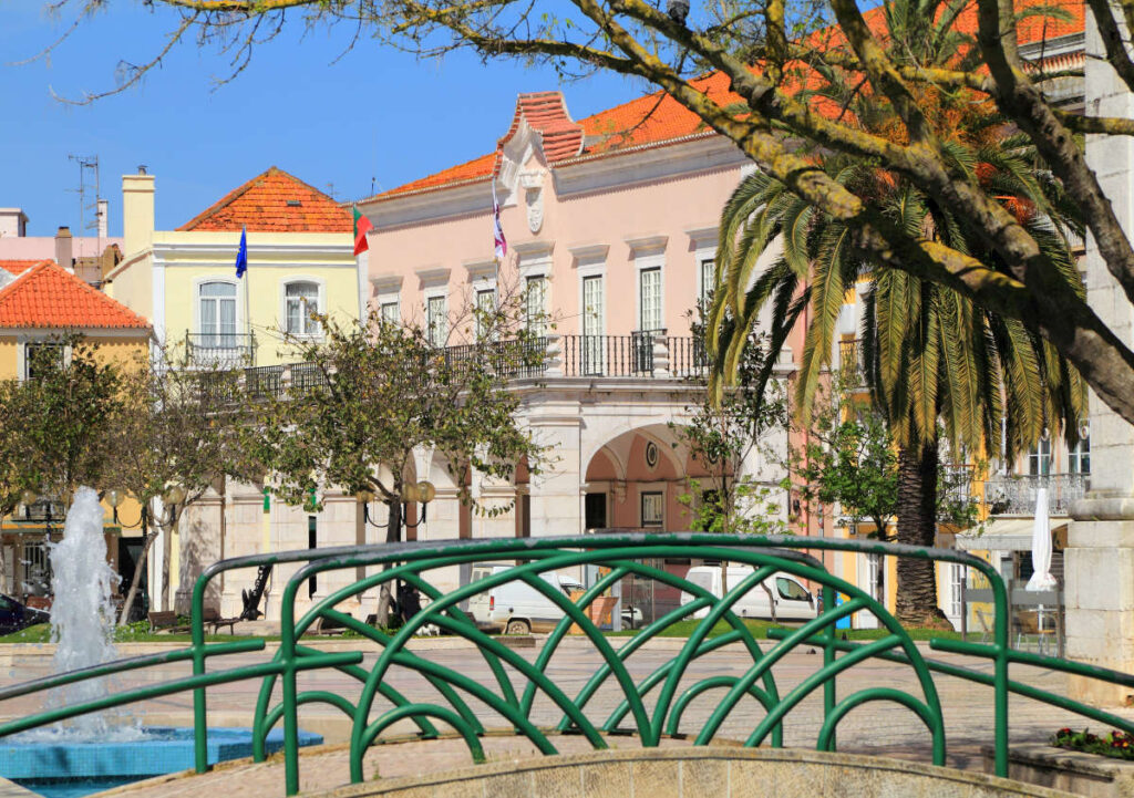 A small green bridge crosses a fountain in a plaza with palm trees and pastel-colored buildings, capturing the charm of coastal towns for late summer.