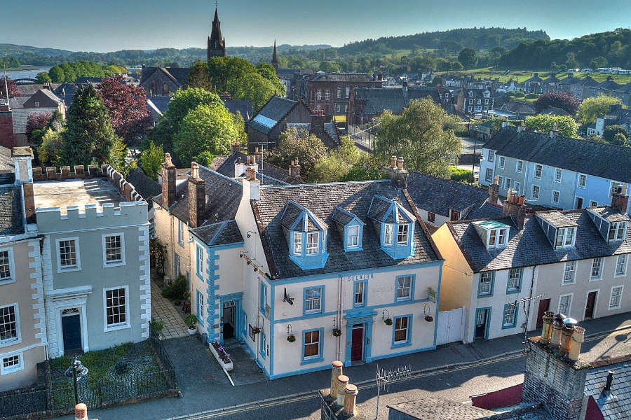 Aerial view of a small town featuring rows of colorful historic buildings, including the charming Selkirk Arms, with trees and a church steeple in the background under a clear blue sky.