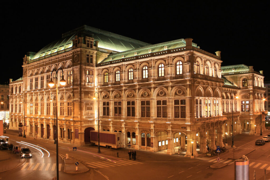 The Opera House in Vienna, a historic building illuminated at night, featuring arched windows and ornate architectural details on a city street corner.