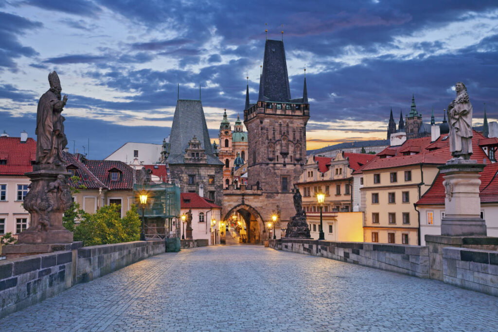 View of Charles Bridge in Prague at dusk, featuring cobblestone walkway, historic statues, and the Old Town Bridge Tower under a cloudy sky.